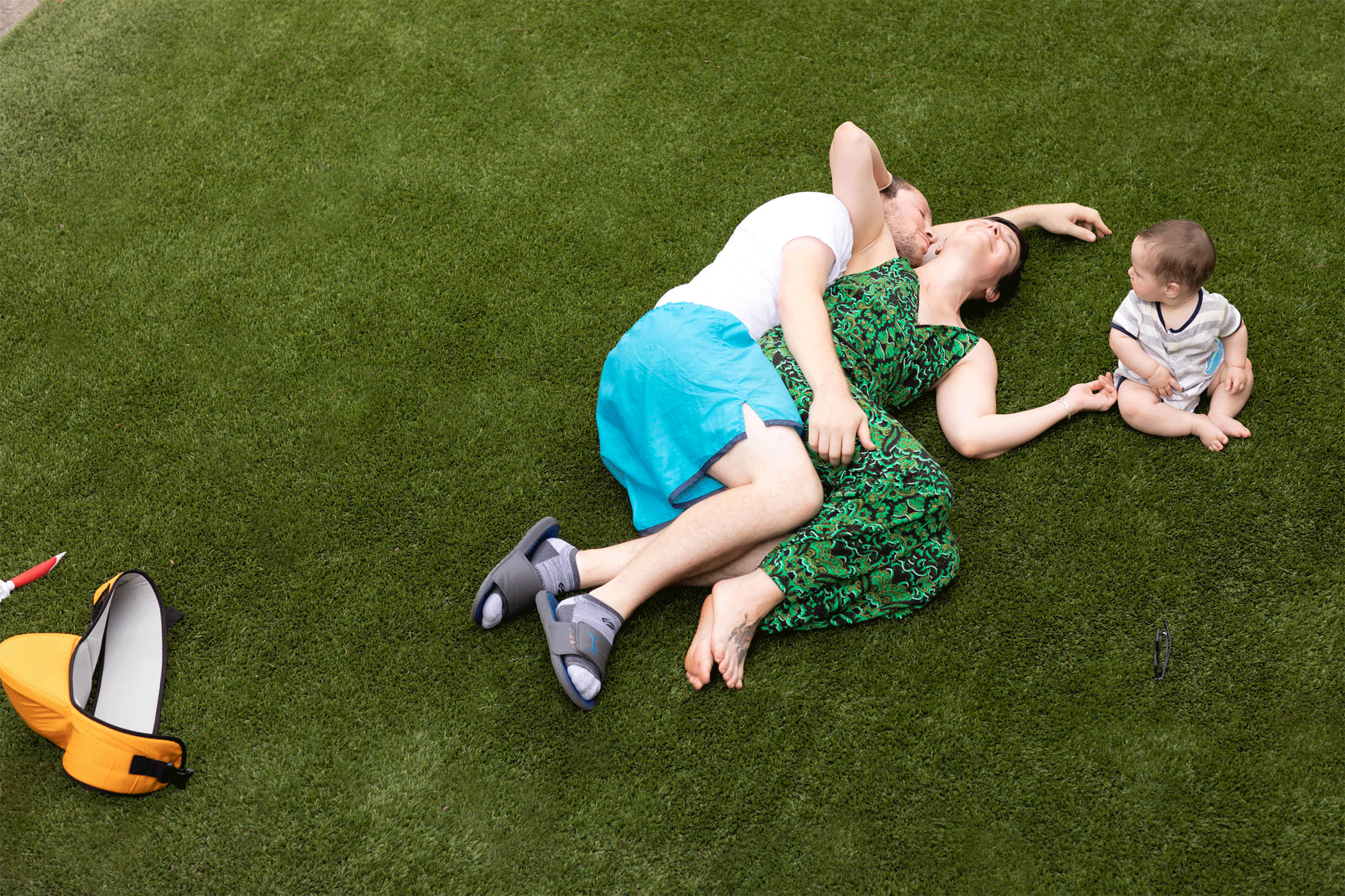 A couple kisses while laying on an astroturf lawn as their 8-month-old son looks on