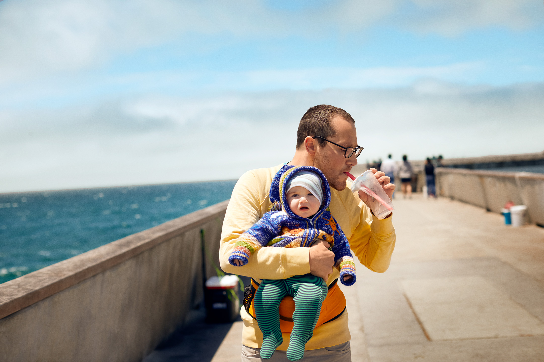 A man drinks boba on a boardwalk by the ocean while holding his infant son