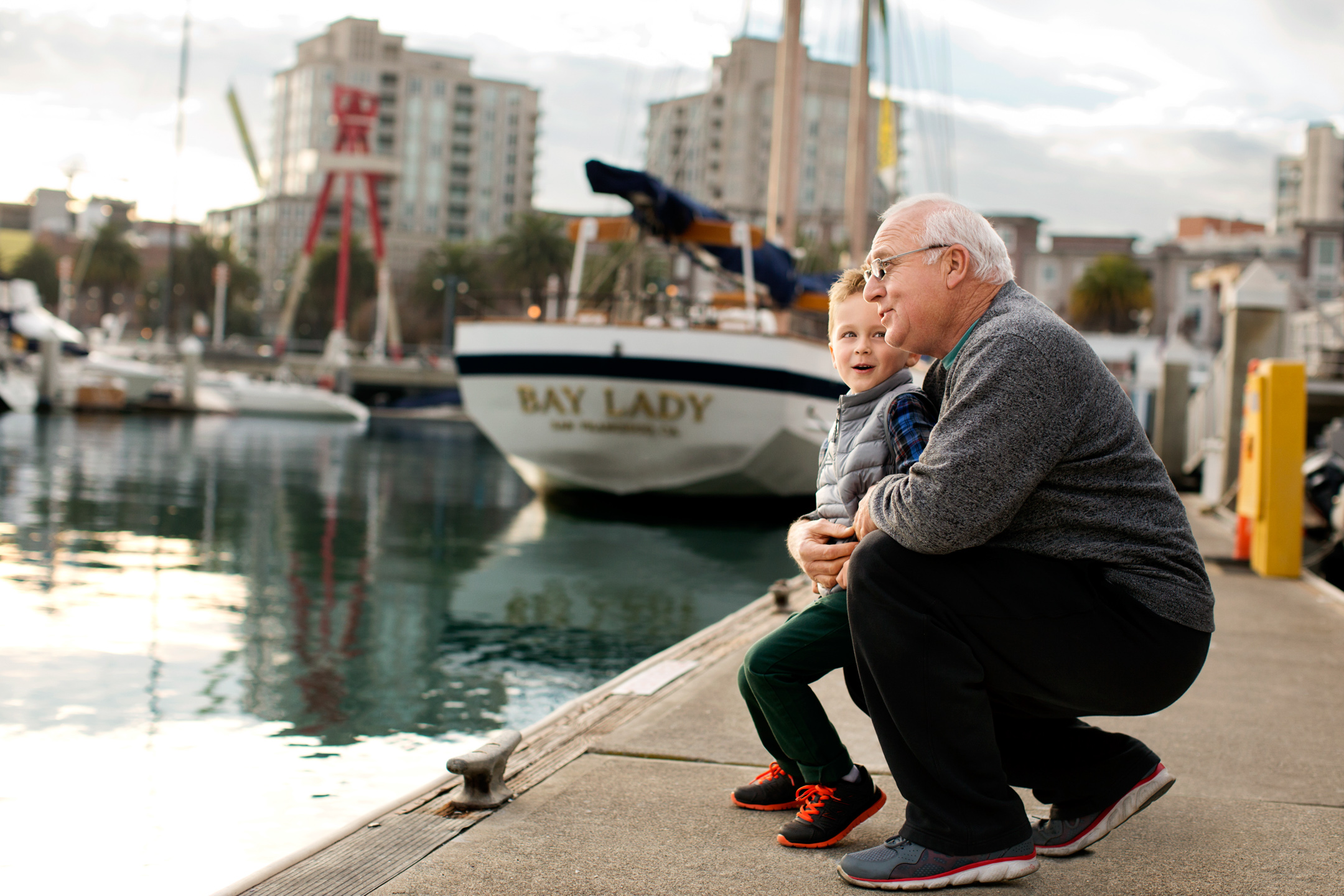 A grandfather holds his grandson as the two look out at a harbor full of boats from a dock