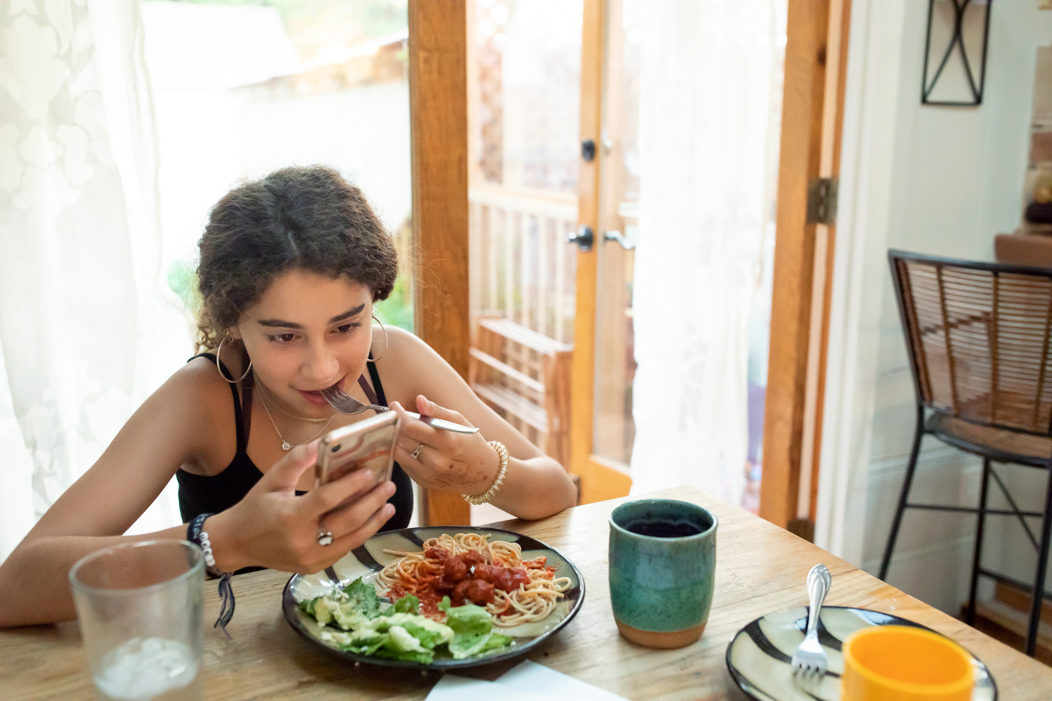 A teen girl eats alone at a table while looking at her phone