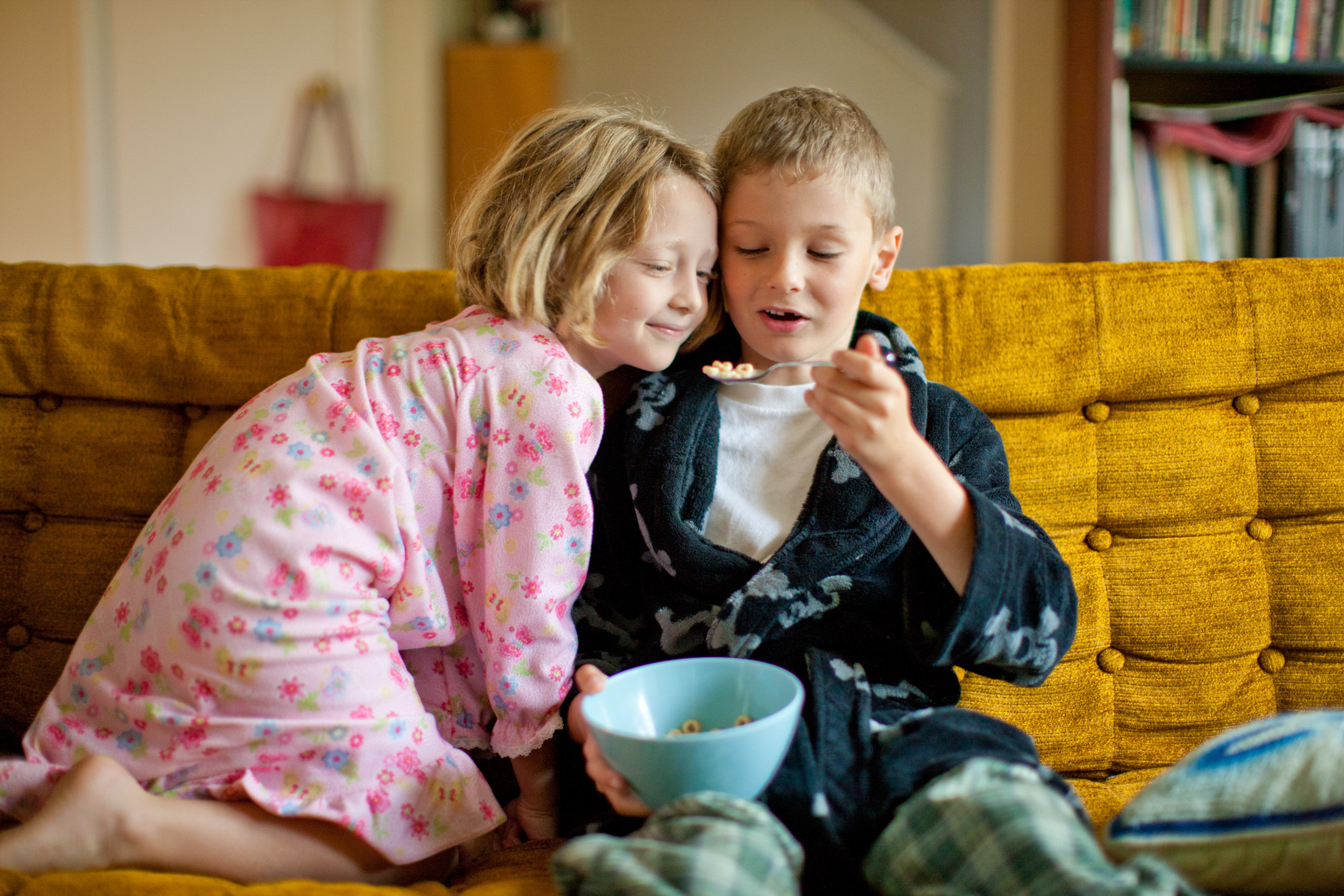 A boy and his sister eat cereal and milk together while sitting on a sofa